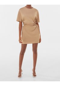 Beżowa sukienka z krótkim rękawem, z bocznym marszczeniem i sznurkami. Wykonana z miękkiego materiału, do kolan, połączona z przezroczystymi sandałami na obcasie.