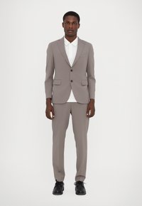 Hellgrauer Anzug aus glattem Stoff, mit einem Zweiknopf-Blazer mit Taschen und schmal geschnittenen Hosen. Kombiniert mit einem weißen Hemd und schwarzen Schuhen.