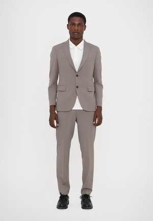 Costume gris clair en tissu lisse, avec un blazer à deux boutons, des poches et un pantalon slim-fit. Associé à une chemise blanche et des chaussures noires.
