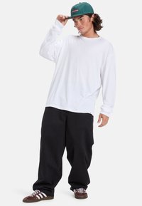 Langärmliges weißes Baumwollhemd, entspannte Passform, kombiniert mit schwarzen lockeren Hosen und bordeauxfarbenen Sneakers mit weißen Streifen; grüße Mütze mit Logo.