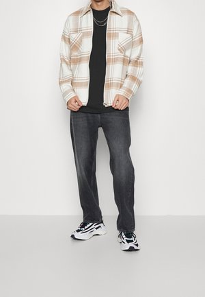 Man draagt een beige-wit geruit jasje met rits over een zwart shirt, donkergrijze spijkerbroek, witte en zwarte sneakers en zilveren kettingkettingen.