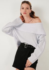Vaaleanharmaa ribattuja neule, jossa on pitkät hihat ja off-the-shoulder-tyyli, yhdistetty mustaan korkeavyötäröiseen hameeseen, jossa on vyö ja hopeinen solki.