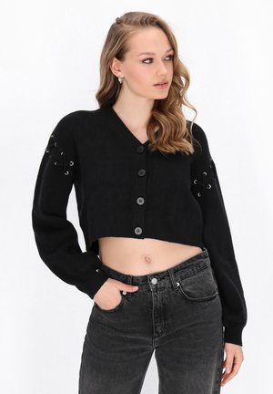 Sort kort cardigan med v-udskæring, fem knapper og snøre detaljer på ærmerne. Båret med højtaljede sorte jeans.