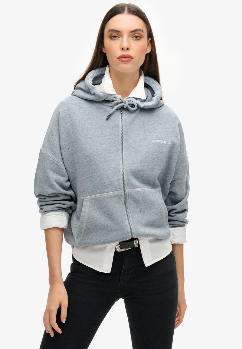 Hellgraue Zip-Hoodie mit einer Kapuze mit Kordelzug, Fronttaschen und gesticktem Logo. Getragen über einem weißen Hemd mit Kragen und schwarzen Hosen.