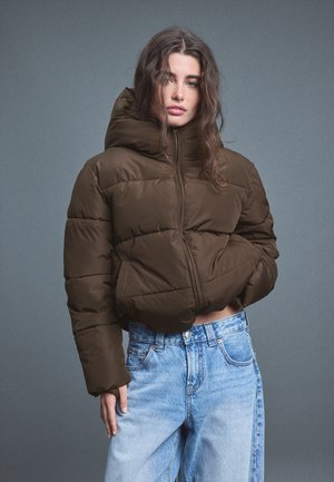 Jeune femme aux longs cheveux ondulés porte une doudoune marron et un jean bleu clair, debout devant un fond gris uni.