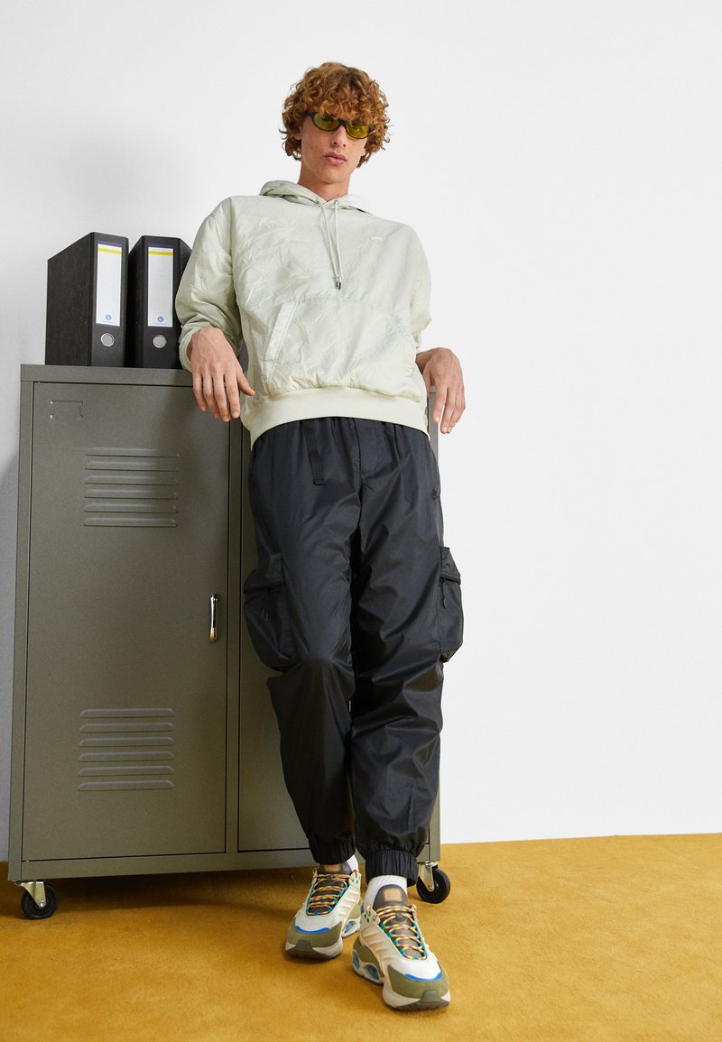 Hellgrüne Kapuzenjacke mit strukturiertem Finish, schwarze Cargo-Hosen und mehrfarbige Sneakers. Steht neben einem grauen Aktenschrank.