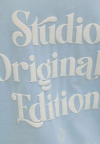 Texte blanc en relief « Studio Original Edition » sur tissu bleu clair, mettant en valeur une police audacieuse de style vintage.