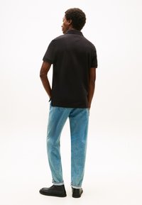 Uomo di spalle che indossa una polo nera a maniche corte, jeans azzurri e scarpe nere, su uno sfondo bianco uniforme.