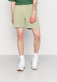 Groene badstof shorts met een elastische tailleband, een gladde textuur en een knielengte, gecombineerd met witte sneakers en zichtbare been tattoos.