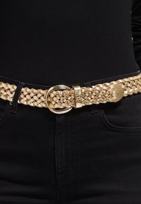 Vævet guld læderbælte med en rund guldspænde, fastgjort gennem en løkke, båret over sort stof.