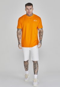 Orange bomulds t-shirt med korte ærmer og rund halsudskæring, parret med hvide shorts. Bemærkelsesværdige tatoveringer på arme og ben; fodtøjet er hvide sneakers.