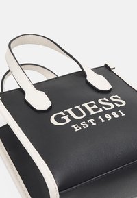 Sac à main en similicuir noir avec des accents blancs. Comprend deux poignées supérieures et le texte "GUESS EST 1981" affiché en blanc de manière proéminente.