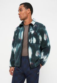 Fleece jacket in teal, black, and white tie-dye pattern with a zippered front, stand collar, and chest pocket. Worn with navy plaid pants.