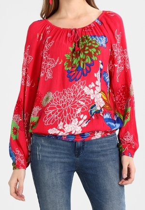 Frau in roter Bluse mit langen Ärmeln, verziert mit bunten Blumen- und Vogelmustern, kombiniert mit blauen Jeans, steht mit entspannten Armen.