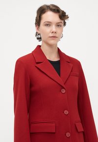Young person with short curly hair wears a red buttoned wool coat over a black top against a plain white background.