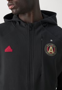 Svart zip-up hoodie med röd Adidas-logotyp, Atlanta United FC-emblem och en texturerad tyg. Inkluderar justerbar huva och dragkedja.