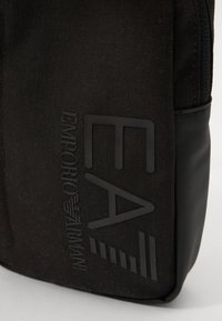 Black fabric bag features a smooth texture, zip closure, and embossed black logo. Side has a contrasting matte finish. Compact design.