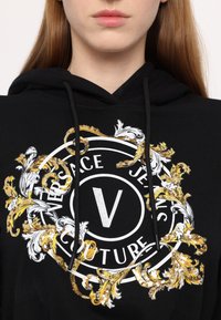 Crna dukserica s centralnim bijelim i zlatnim logotipom Versace Jeans Couture, ukrašenim bogatim zlatnim cvjetnim detaljima i teksturiranim dizajnom.