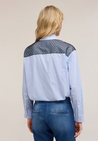 Camicetta a righe blu con schiena in rete nera trasparente, caratterizzata da un motivo texturizzato sulla rete e maniche lunghe, abbinata a jeans blu.