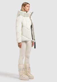 Hvid quiltet pufferjakke med høj krave, lynlås lukning og sort inderside. Sammen med lyse beige leggings og knæhøje støvler.