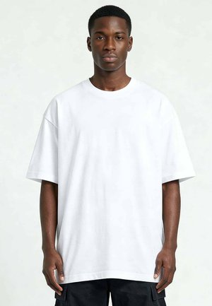 Jonge zwarte man met kort haar, gekleed in een simpele oversized witte T-shirt en zwarte broek, staand tegen een lichtgrijze achtergrond.