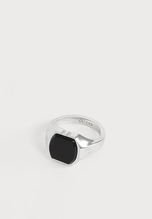 Bague en argent avec une pierre noire rectangulaire au centre et "GUESS" gravé à l'intérieur de la bague sur fond blanc.