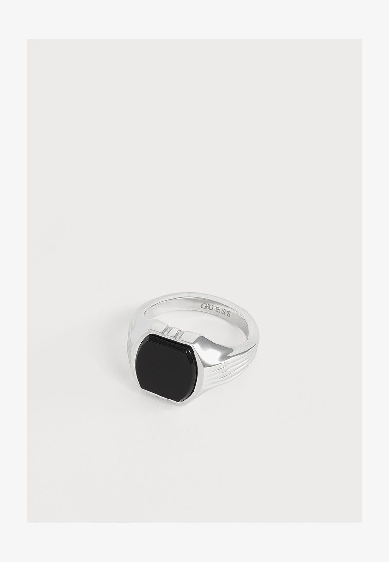 Bague en argent avec une pierre noire rectangulaire au centre et "GUESS" gravé à l'intérieur de la bague sur fond blanc.