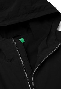 Černá bunda s kapucí, s předním zipem s odrazovými prvky a měkkou fleecovou podšívkou. Uvnitř viditelná zelená štítek.