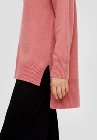 Long pull rose avec poignets et ourlet côtelés, présentant des fentes latérales. Texture douce, coupe décontractée, associé à un bas noir.