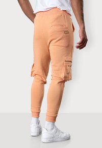 Die Pfirsich-farbenen Cargo-Jogginghosen verfügen über einen gerippten Bund, eine Slim-Fit-Passform, Reißverschlusstaschen an den Seiten und ein gesticktes Logo-Detail, kombiniert mit weißen Sneakers.