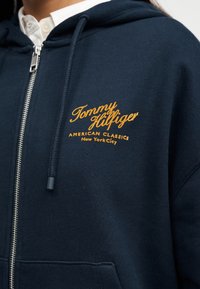 Μπλε ναυτικό φούτερ με φερμουάρ και κορδόνια, με κίτρινη επιγραφή "Tommy Hilfiger American Classics New York City" στο στήθος, φορεμένο πάνω από λευκό πουκάμισο με γιακά.