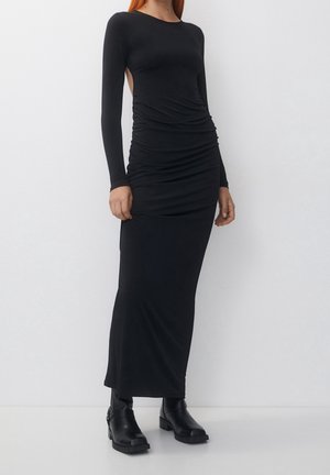 Vrouw draagt een lange zwarte nauwsluitende jurk met lange mouwen en zijopeningen, gecombineerd met zwarte enkellaarzen, staand tegen een effen witte muur.