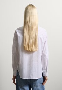 Camicia bianca a maniche lunghe con righe verticali, polsini a bottone e vestibilità rilassata. Indossata con jeans blu a vita alta. Tessuto con una consistenza liscia.