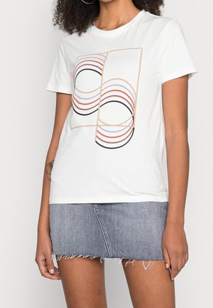 Kvinna i en vit t-shirt med abstracta kurviga linjemönster, matchad med en ljusblå denim mini-short med fransar.