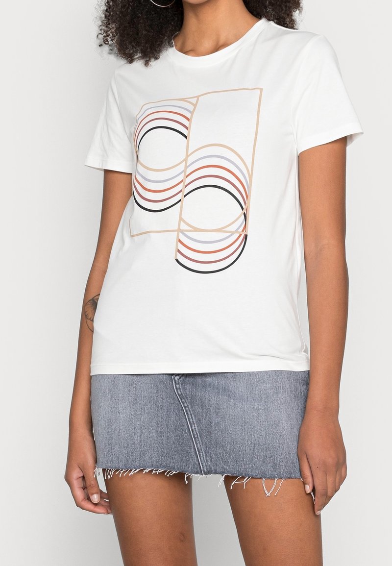 Žena nosící bílé tričko s abstraktními křivkami v designu, které je doplněno světle modrou denimovou mini sukní s třepeným lemem.