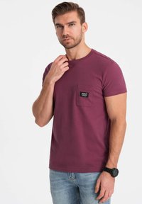 Mørk bordeaux t-shirt i bomuld med rund hals, korte ærmer og en brystlomme med et mærke; iført lyseblå jeans.
