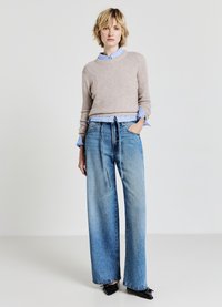 Beiges Strickpullover über einem hellblauen Kragenhemd, kombiniert mit lockeren, hellblauen Jeanshosen. Das Outfit umfasst schwarze Schuhe.