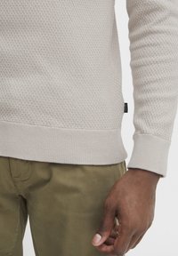 Beige textúrált pulóver bordázott nyakkal és mandzsettákkal, olívazöld nadrággal párosítva. Diszkrét márkajelzés található az oldalon.