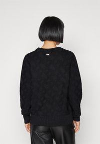 Jersey negro de punto con un patrón texturizado y puños acanalados, con un logo sutil en la parte trasera del escote. Tejido suave y corte relajado.
