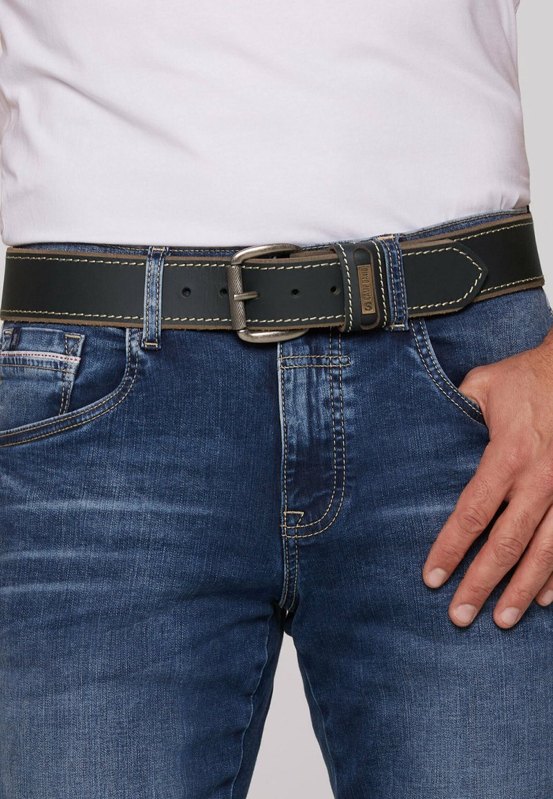 Schwarzer Ledergürtel mit einer silbernen Metallschnalle, verziert mit kontrastierendem beigem Näht. Getragen über blauen Jeans.