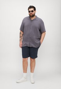 Kortermet, knappet skjorte i lys grå, kombinert med marineblå stripete shorts. Modellen har på seg hvite joggesko og solbriller, med lyse sokker.