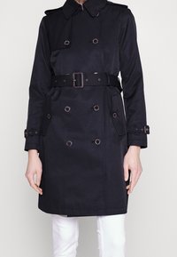 Mörkblå trench coat med dubbelknäppt design, stor krage, bälte vid midjan och knappramar. Tillverkad av mjukt tyg, med två sidofickor.