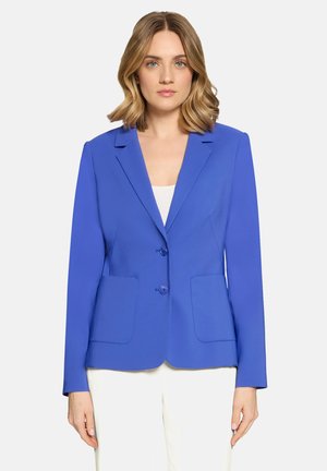 Kvinde med skulderlangt blondt hår iført en tætsiddende, lys blå blazer over en hvid top og hvide bukser foran en ensartet baggrund.