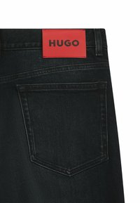 Mustat farkut, joissa on punainen nahkapatentti, jossa lukee "HUGO". Takana on tasku ja kontrastivärinen ompelu saumoissa.