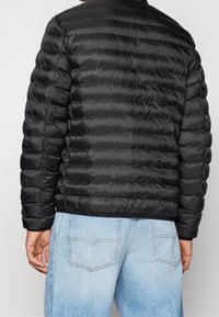 Musta puffer-takki, jossa on vaakasuorat tikatut paneelit, sileä ja kiiltävä pinta sekä suora helma. Päällä vaaleansinisten farkkujen päällä.