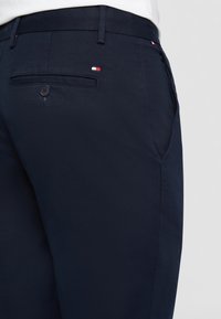 Close-up van marineblauwe broek met een achterzak met knoop en kleine Tommy Hilfiger vlaggetjes bij de zak en tailleband.
