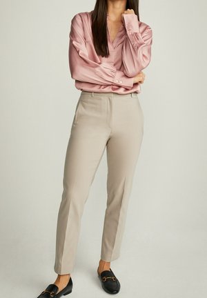 Woman wearing a pink satin blouse, beige tailored pants, and black loafers, standing with arms crossed against a plain background.