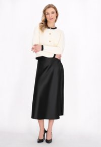 Cardigan en tricot ivoire avec bordure noire et boutons dorés, associé à une jupe midi en satin noir. Le modèle porte des escarpins noirs.
