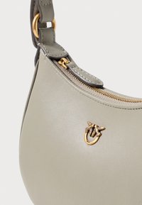 Khaki kožená kabelka s zakriveným siluetou, zlatými zipsami a dekoračným kovovým akcentom s prepletenými listami.