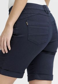 Pantalones cortos de mezclilla azul marino con dobladillo vuelto, dos bolsillos traseros y un detalle en dorado en el bolsillo. Textura suave y diseño ajustado.
